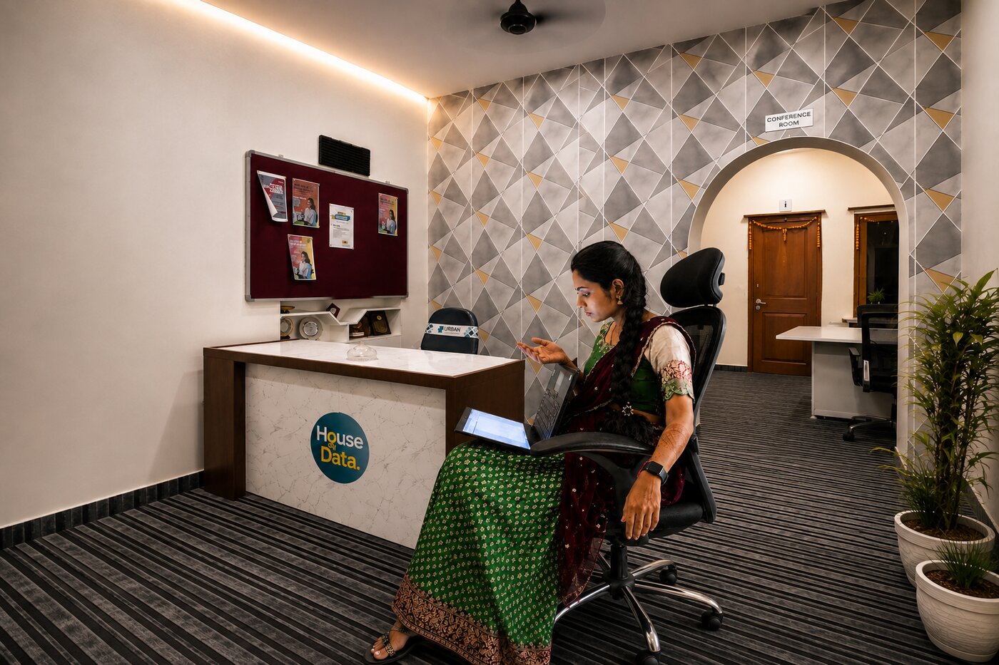 Vinitha at the House of Data office lounge, working on a laptop, with marigold torans hung over the archway behind her.